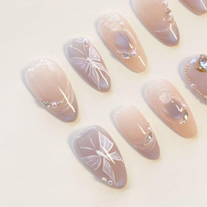 A set of almond-shaped press-on nails featuring a soft nude gradient base. The designs include delicate white hand-painted butterfly wings, small iridescent rhinestones, and tiny white pearls arranged in elegant patterns across the nails.