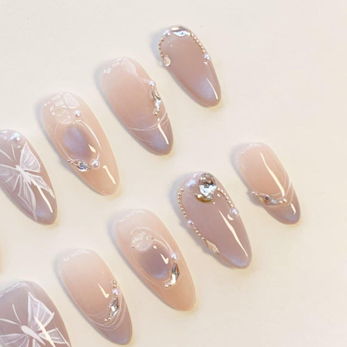 A set of almond-shaped press-on nails featuring a soft nude to mauve gradient. The nails are decorated with white hand-painted butterfly wing designs, small clear rhinestones, and tiny white pearls arranged in delicate patterns.