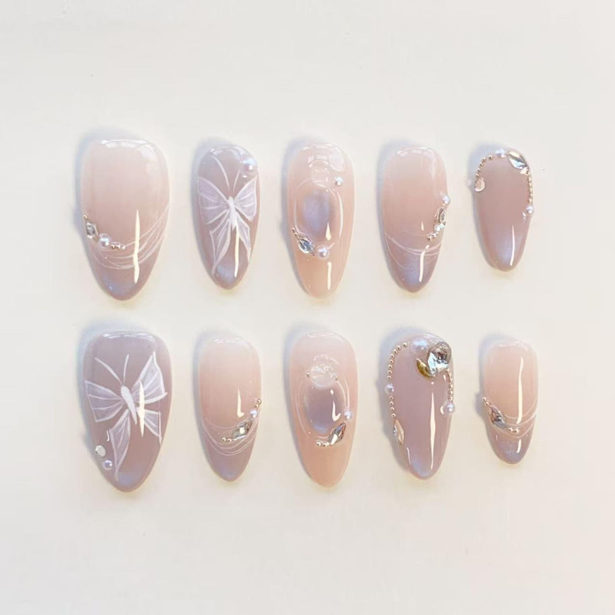 A set of ten almond-shaped press-on nails in a soft nude and mauve gradient. The design features hand-painted white butterfly motifs, delicate silver chains, small clear rhinestones, and tiny white pearls arranged on the nail surfaces.