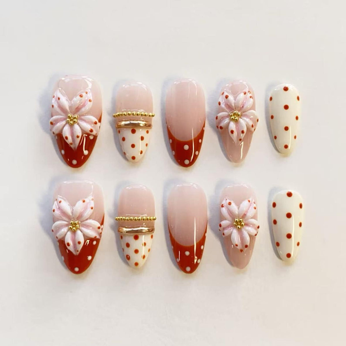 A set of ten almond-shaped press-on nails featuring a nude base with various designs including raised white and pink floral accents, red polka dots on white backgrounds, gold metallic bands, and red French tips with white polka dots.