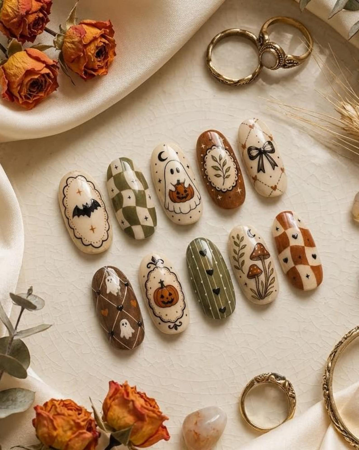 A set of ten oval-shaped press-on nails featuring various autumn and Halloween-themed designs, including ghosts, pumpkins, bats, mushrooms, bows, and checkered patterns in shades of cream, olive green, and burnt orange.