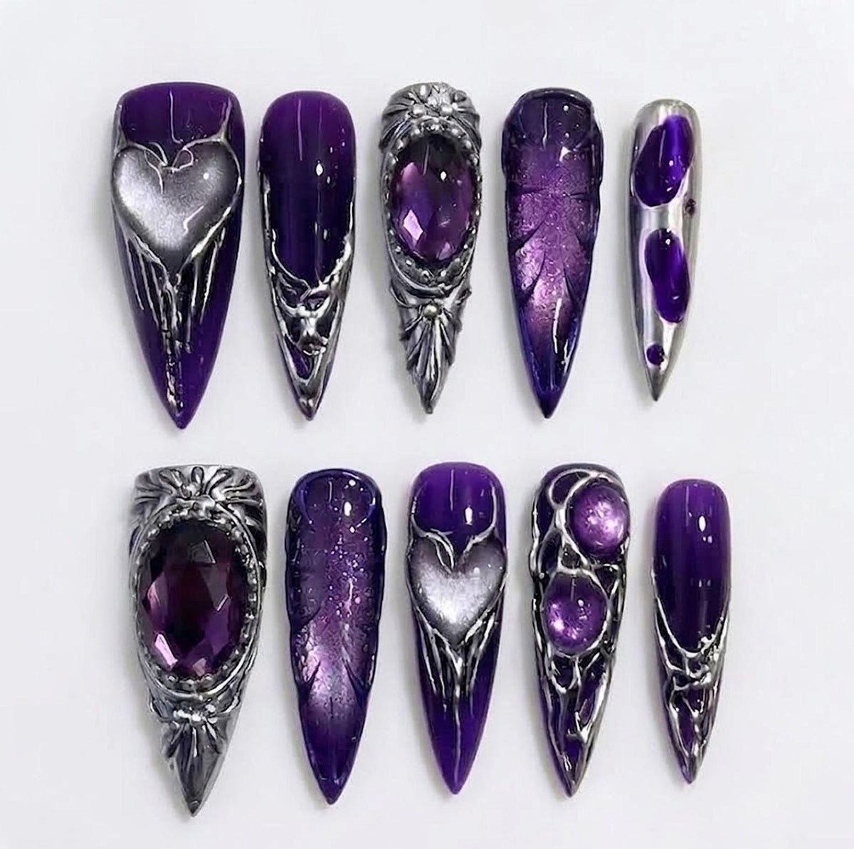 A set of ten stiletto-shaped press-on nails in deep purple with metallic silver accents. Designs include raised silver heart motifs, large purple faceted gems, intricate silver filigree, textured leaf patterns, and glossy purple finishes.