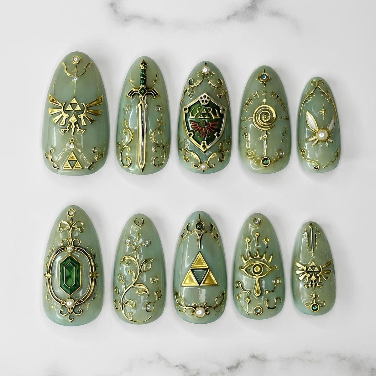 A set of ten almond-shaped press-on nails in a sage green hue, featuring intricate gold-toned metallic embellishments including a sword, a shield, a triforce symbol, a fairy, a green gemstone, and an eye motif with small pearl accents.