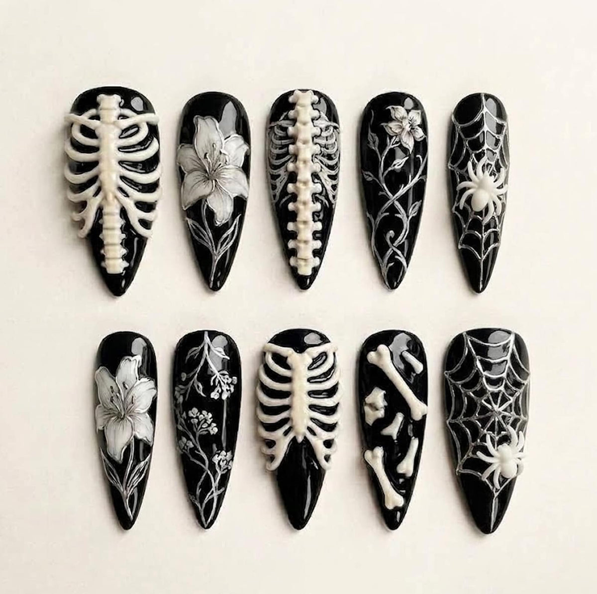 A set of ten almond-shaped press-on nails with a glossy black base. The designs feature white raised 3D details including human rib cages, spines, lilies, intricate floral vines, spider webs with spiders, and scattered bone fragments.