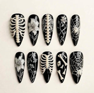A set of ten almond-shaped press-on nails with a glossy black base. The designs feature white raised 3D details including human rib cages, spines, lilies, intricate floral vines, spider webs with spiders, and scattered bone fragments.