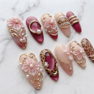 A set of ten almond-shaped press-on nails featuring a mix of designs including 3D pink flowers, gold filigree, white pearls, leopard print patterns, and deep burgundy cat-eye polish with gold metal accents and small hanging chains.