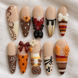 A set of ten almond-shaped press-on nails featuring various designs including brown swirls, polka dots, a black cat, piano keys, a white bow, a dark red bow, a white flower, a brown bear, musical notes, and autumn leaves.