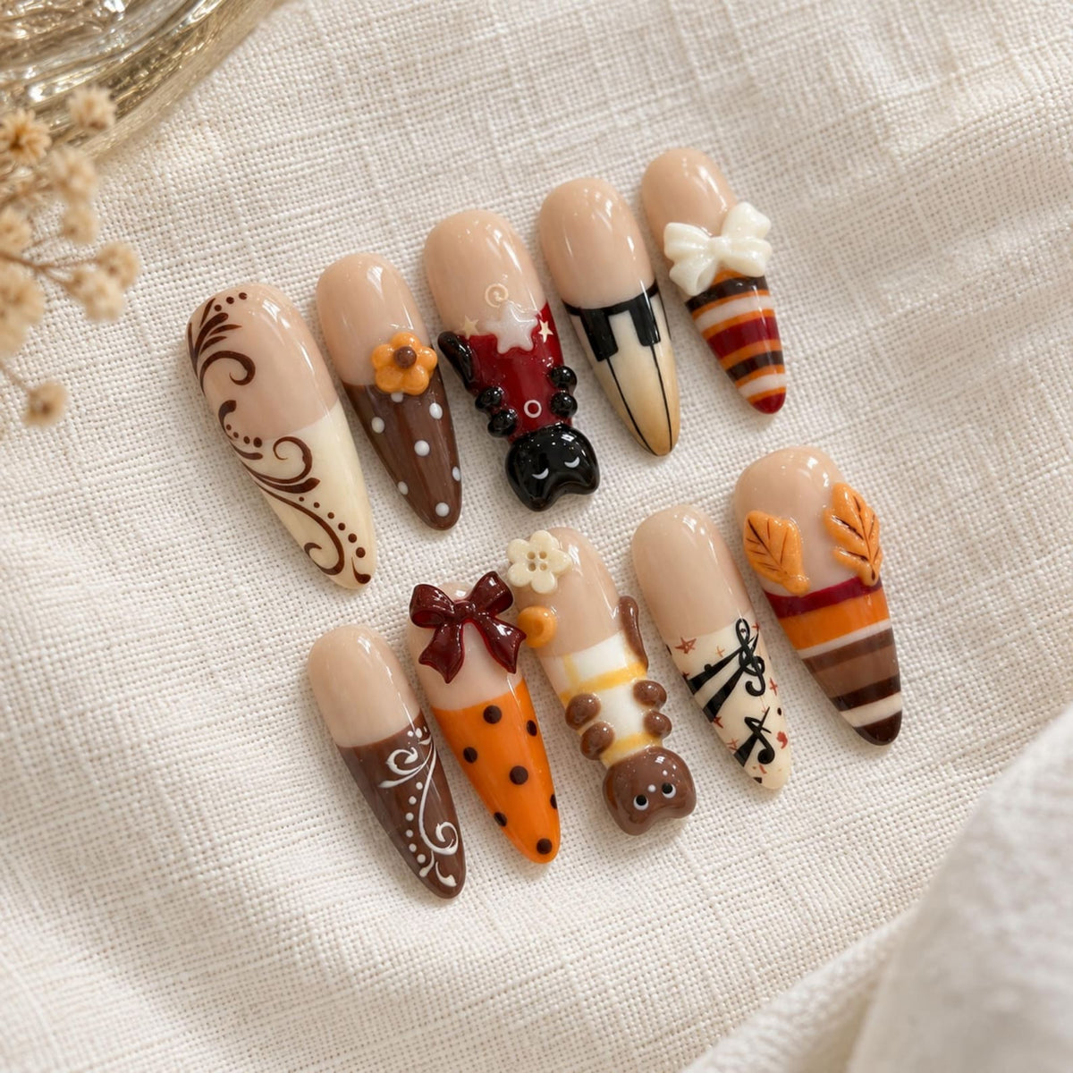 A set of ten almond-shaped press-on nails featuring a nude base with various decorative elements including brown swirls, orange polka dots, a black cat, piano keys, a white bow, musical notes, autumn leaves, and striped patterns.