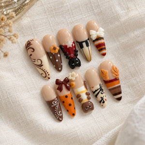 A set of ten almond-shaped press-on nails featuring a nude base with various decorative elements including brown swirls, orange polka dots, a black cat, piano keys, a white bow, musical notes, autumn leaves, and striped patterns.