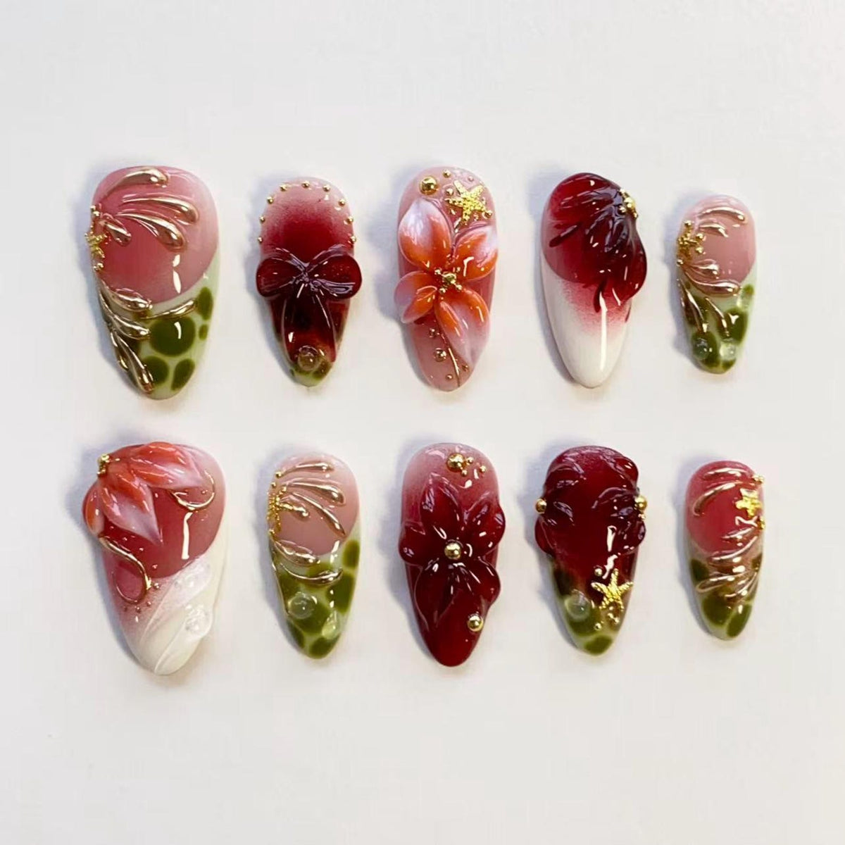 A set of ten press-on nails featuring a variety of designs in shades of deep red, pink, white, and olive green. The nails are decorated with three-dimensional floral motifs, gold metallic accents, and small gold beads.