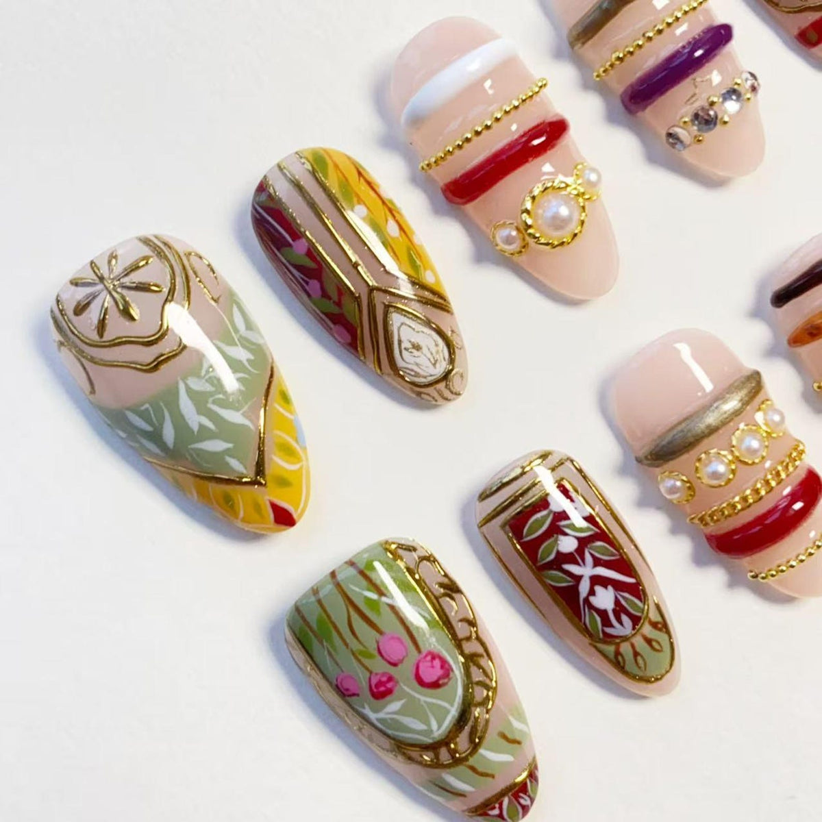 A set of almond-shaped press-on nails featuring a nude base with intricate designs. Decorations include gold metallic lines, white and green leaf patterns, small pink floral accents, red glossy stripes, white pearls, and gold chain details.