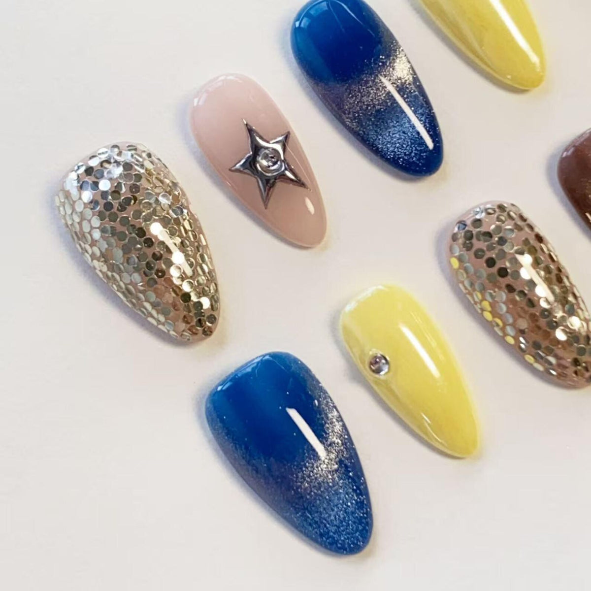 A set of almond-shaped press-on nails featuring various designs including gold hexagonal glitter, a nude base with a silver star charm, deep blue gradient with shimmer, and bright yellow with a single clear rhinestone accent.