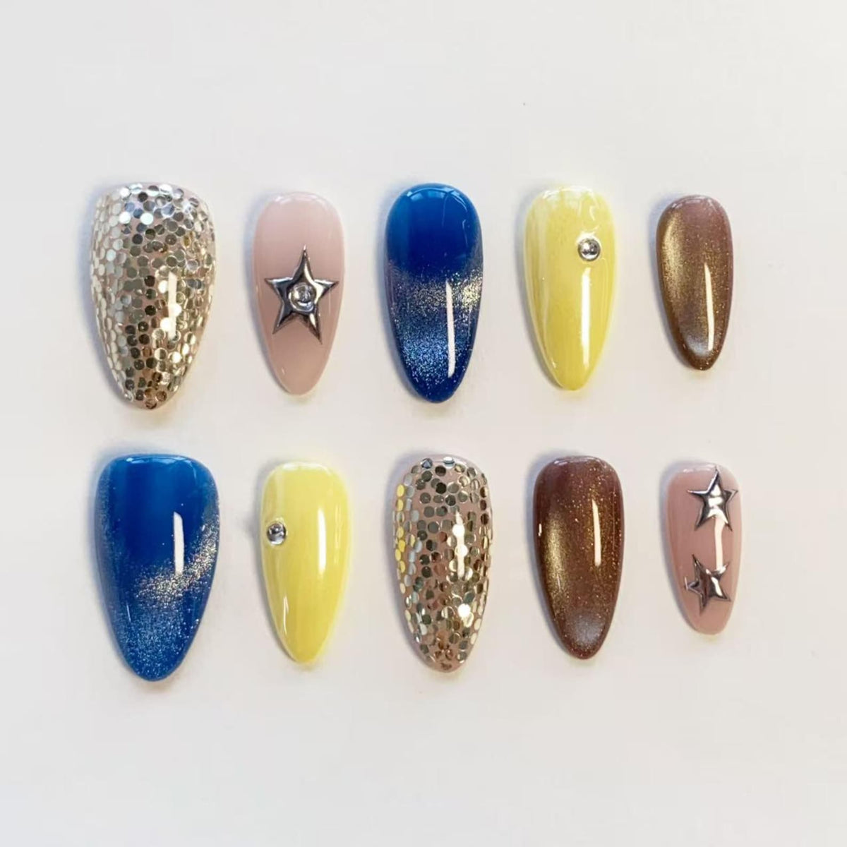 A set of ten press-on nails featuring various designs including silver glitter, solid yellow with a single rhinestone, deep blue with a glitter gradient, metallic brown, and light pink with silver star charms and a single star cutout.