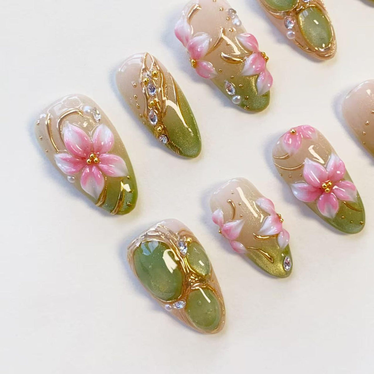 A set of press-on nails featuring a gradient of soft beige to olive green. The designs include three-dimensional pink floral accents, gold metallic line detailing, small clear rhinestones, and smooth green cabochon-like embellishments.