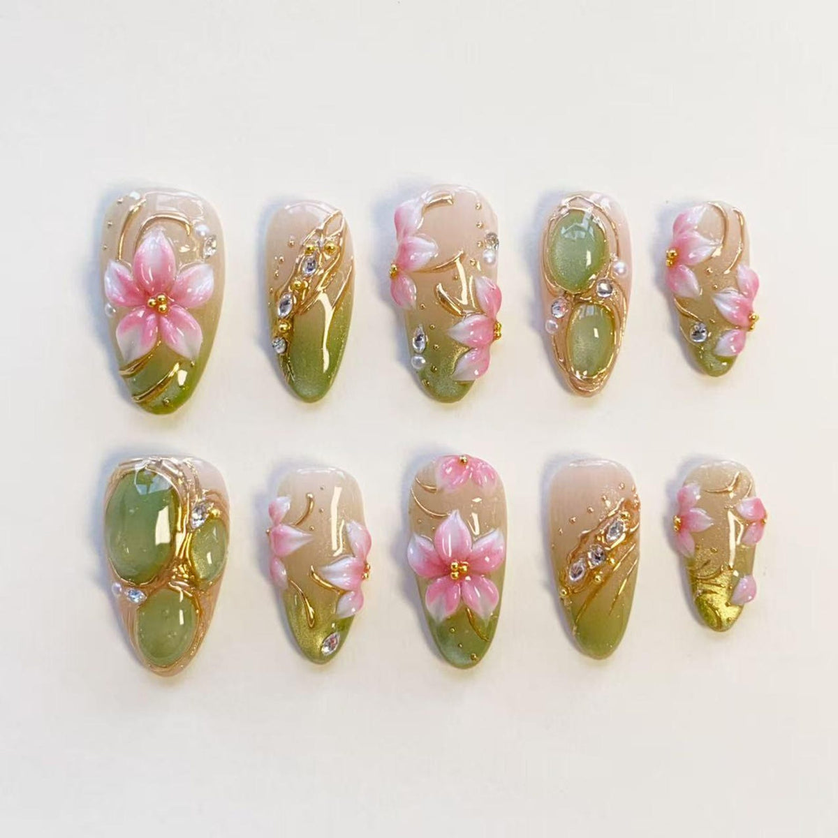 A set of ten press-on nails featuring a gradient of soft beige to sage green. The designs include raised pink floral motifs, gold metallic accents, small clear rhinestones, and smooth green oval cabochons arranged in various patterns.