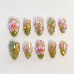 A set of ten press-on nails featuring a gradient of soft beige to sage green. The designs include raised pink floral motifs, gold metallic accents, small clear rhinestones, and smooth green oval cabochons arranged in various patterns.