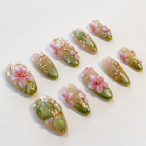 A set of ten press-on nails featuring a gradient design transitioning from beige to olive green. Each nail is decorated with three-dimensional pink floral accents, gold metallic detailing, and small clear rhinestones.