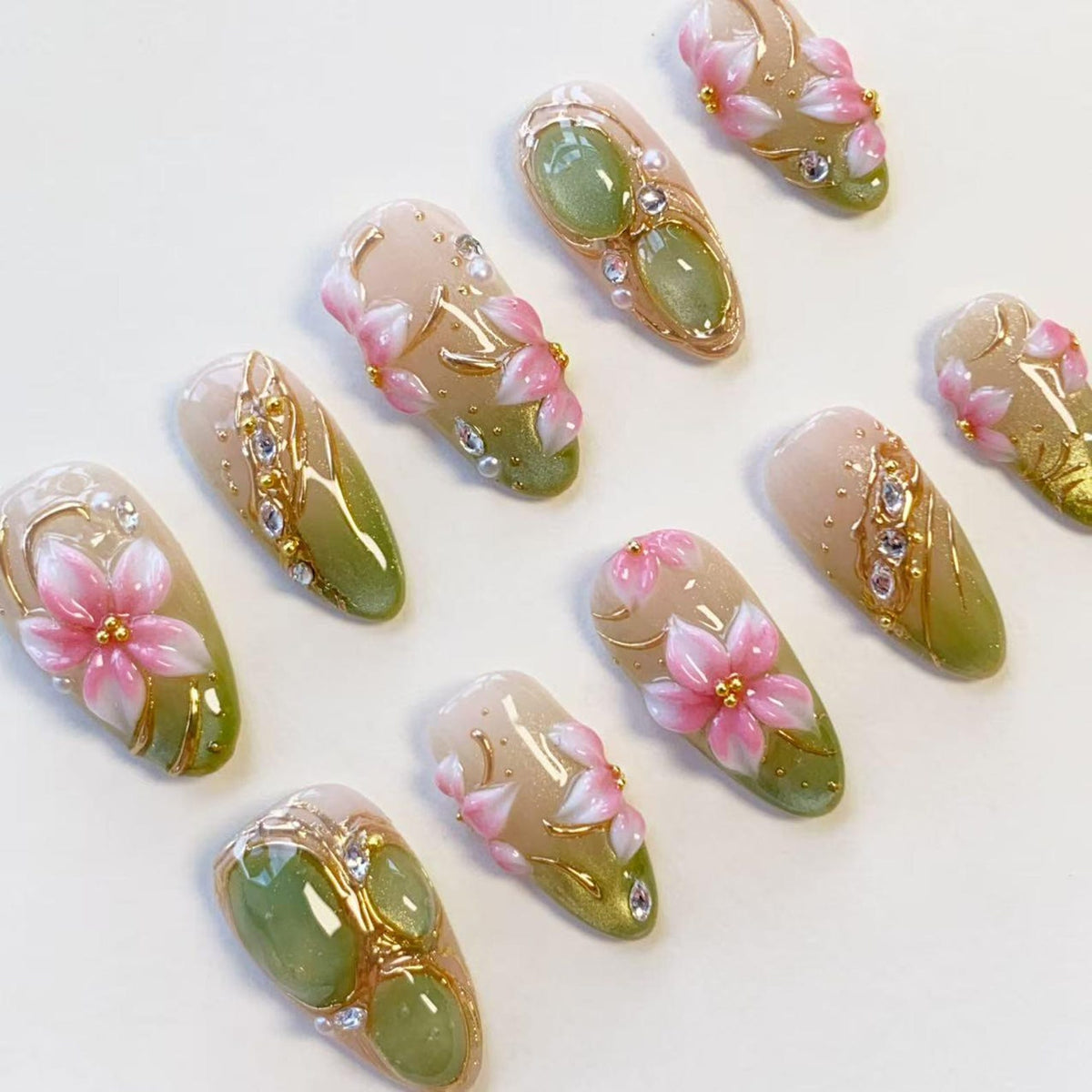 A set of ten almond-shaped press-on nails featuring a gradient design transitioning from a nude base to a sage green tip. The nails are decorated with hand-painted pink flowers, gold metallic accents, small clear rhinestones, and pearls.