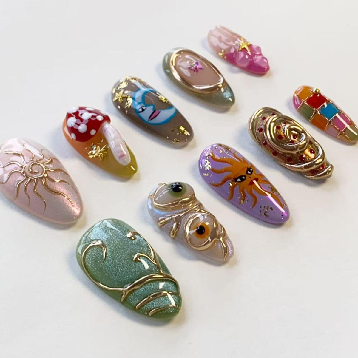 A collection of ten almond-shaped press-on nails featuring diverse designs including a red mushroom, a crescent moon, a sun face, a green textured surface with gold swirls, and various colorful patterns with gold accents and embellishments.