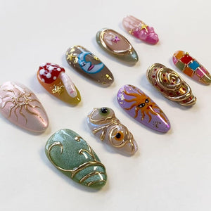 A collection of ten almond-shaped press-on nails featuring diverse designs including a red mushroom, a crescent moon, a sun face, a green textured surface with gold swirls, and various colorful patterns with gold accents and embellishments.