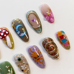 A collection of ten almond-shaped press-on nails featuring diverse designs including a red mushroom, a blue crescent moon, a purple butterfly, a pink starfish, a yellow sun, a gold spiral, and a colorful checkered pattern with gold accents.