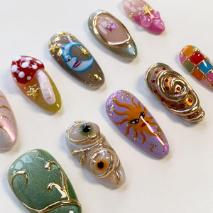 A collection of ten almond-shaped press-on nails featuring diverse designs including a red mushroom, a blue crescent moon, a purple sun, a green cat-eye finish, gold foil accents, and raised gold wire detailing on a white surface.