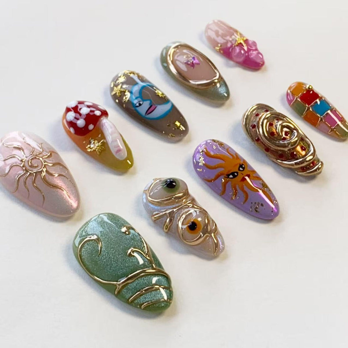 A collection of ten almond-shaped press-on nails featuring diverse designs including a gold sun, a red mushroom, a crescent moon, a purple sun face, gold swirls, and colorful geometric patterns, all accented with gold foil and 3D details.