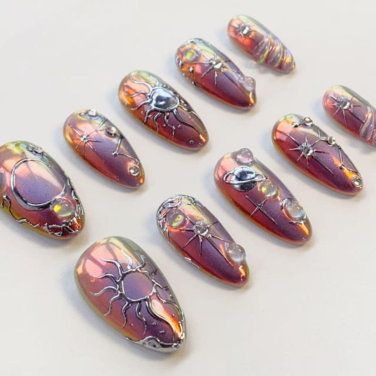 A set of ten almond-shaped press-on nails featuring a metallic, iridescent purple and gold finish. Each nail is decorated with raised silver celestial motifs, including suns, stars, and planets, accented with small, clear, round rhinestones.