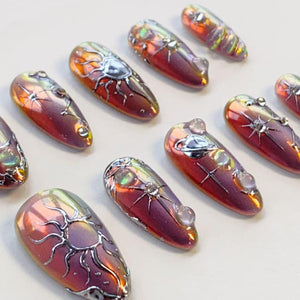 A set of almond-shaped press-on nails featuring a metallic, iridescent red and purple finish. Each nail is decorated with raised silver celestial motifs, including suns, stars, and planets, accented with small, clear, rounded beads.