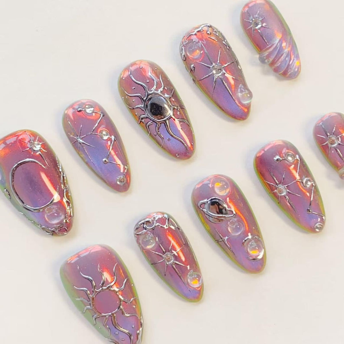 A set of ten almond-shaped press-on nails featuring an iridescent purple and pink finish. Each nail is decorated with raised silver metallic celestial motifs, including suns, moons, stars, and planets, accented with small clear beads.