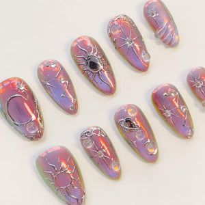 A set of ten almond-shaped press-on nails featuring an iridescent purple and pink finish. Each nail is decorated with raised silver metallic celestial motifs, including suns, moons, stars, and planets, accented with small clear beads.