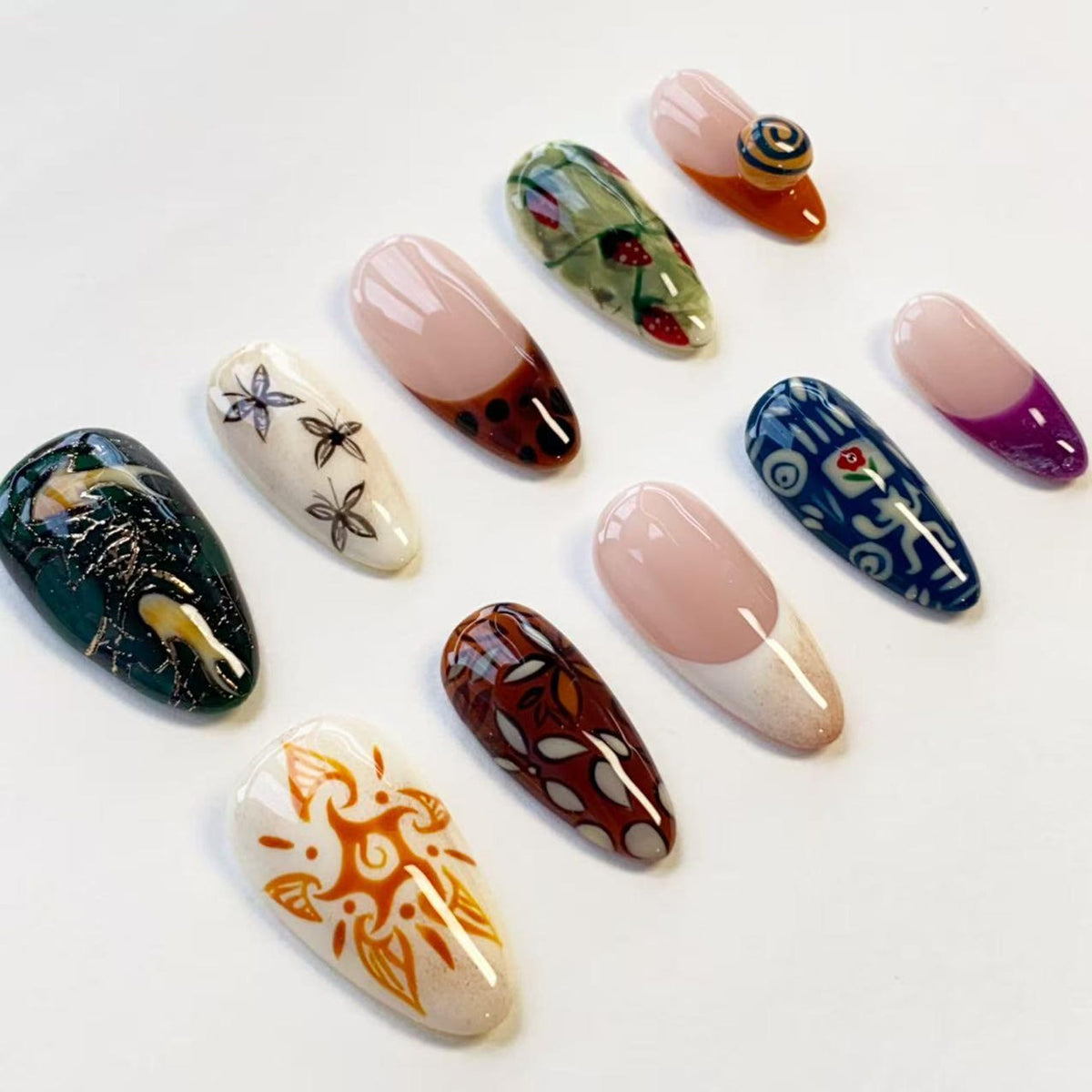 A collection of ten almond-shaped press-on nails featuring diverse designs including a dark green textured pattern, white with black floral sketches, brown tortoiseshell, strawberry motifs, a blue abstract print, and orange sun art.