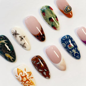 A collection of ten almond-shaped artificial nail tips featuring diverse designs including strawberry patterns, black butterfly illustrations, blue and white geometric motifs, brown leaf prints, and orange sunburst patterns on a white background.