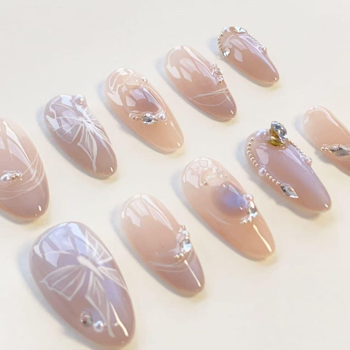 A set of ten almond-shaped press-on nails in a soft nude pink tone. The nails feature white hand-painted ribbon bow designs, small clear rhinestones, and tiny metallic beads arranged in delicate patterns across the glossy surfaces.