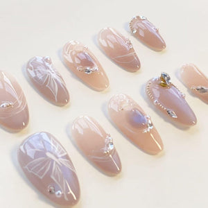 A set of ten almond-shaped press-on nails in a soft nude pink tone. The nails feature white hand-painted ribbon bow designs, small clear rhinestones, and tiny metallic beads arranged in delicate patterns across the glossy surfaces.