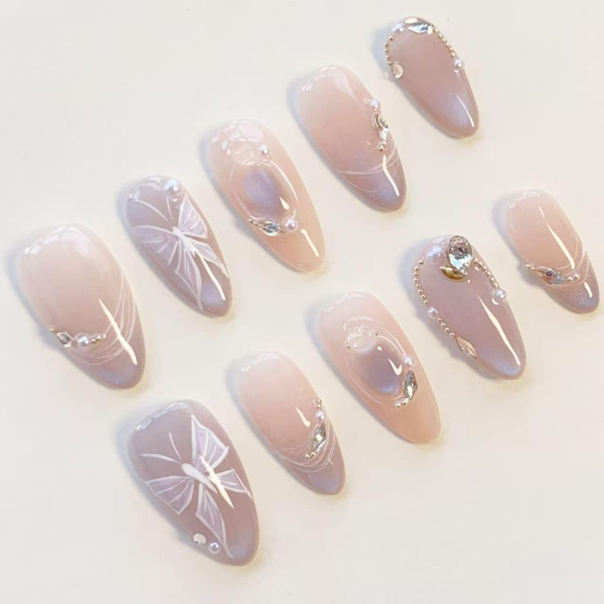 A set of ten almond-shaped press-on nails featuring a soft nude and lavender gradient base. The design includes hand-painted white butterfly wings, small iridescent rhinestones, and tiny white pearls arranged in delicate patterns.
