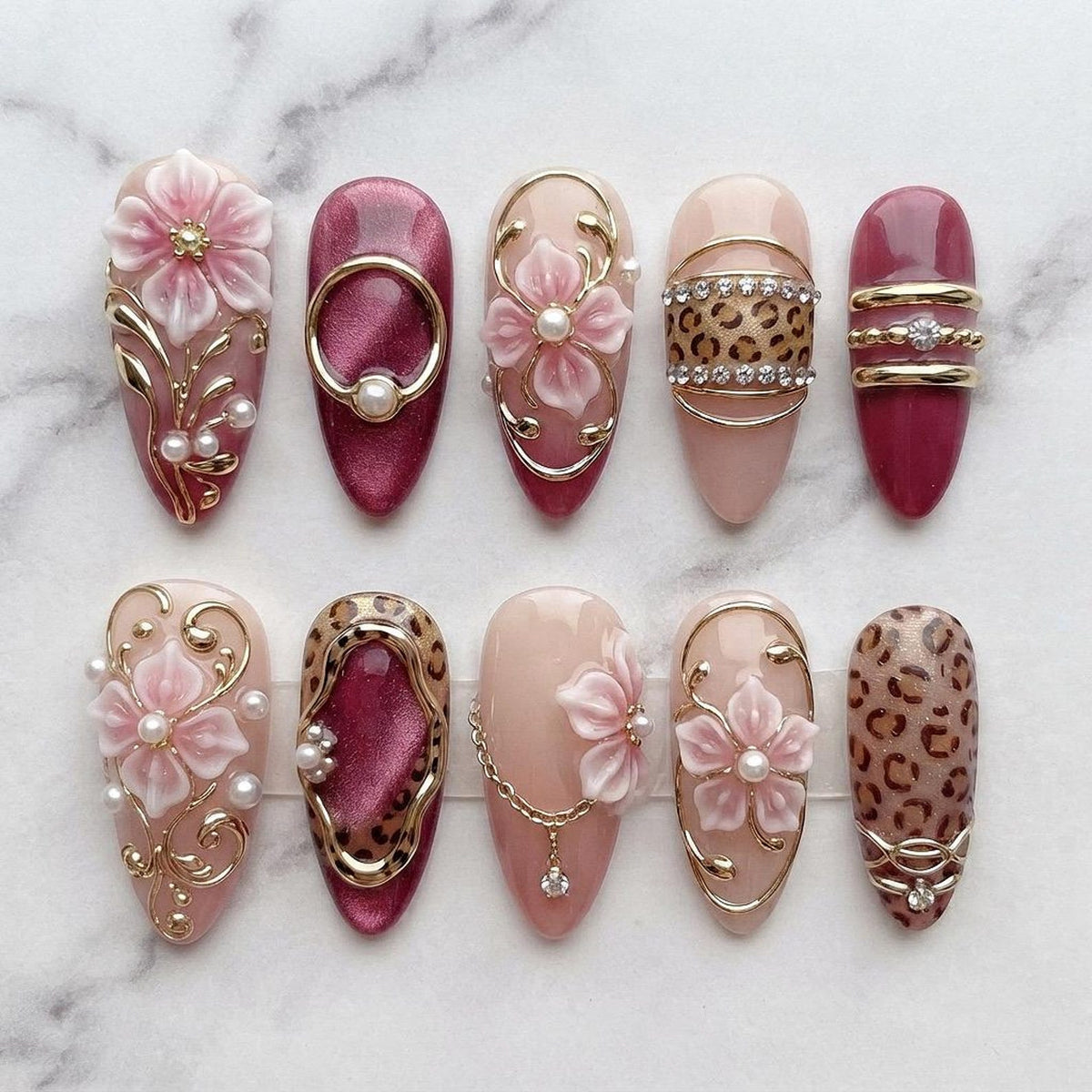A set of ten almond-shaped press-on nails featuring a mix of designs including 3D pink flowers, gold metal accents, white pearls, leopard print patterns, and deep magenta cat-eye polish. Each nail is decorated with intricate gold filigree.
