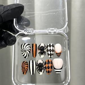A set of ten press-on nails in a clear plastic case, featuring Halloween-themed designs including black and white spirals, orange and black stripes, raised black spider accents, black and white harlequin patterns, and French tips.