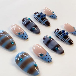 A set of ten almond-shaped press-on nails featuring a mix of designs including light blue and brown horizontal stripes, light blue with black polka dots, and a blue and black plaid pattern, all accented with small blue rhinestones.