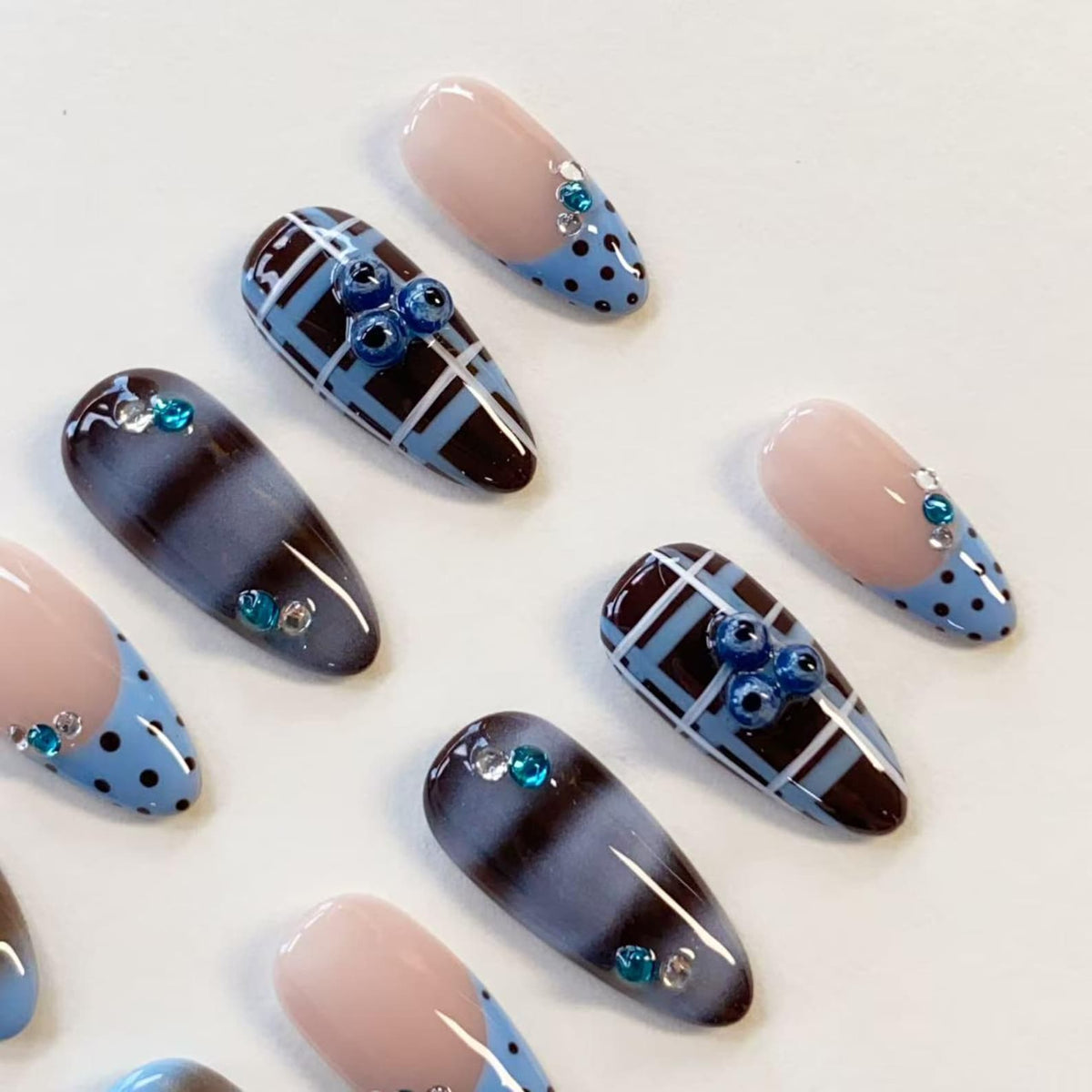 A set of almond-shaped press-on nails featuring a variety of designs including light blue tips with dark blue polka dots, dark brown and light blue plaid patterns, and dark brown ombre gradients, all accented with small blue rhinestones.