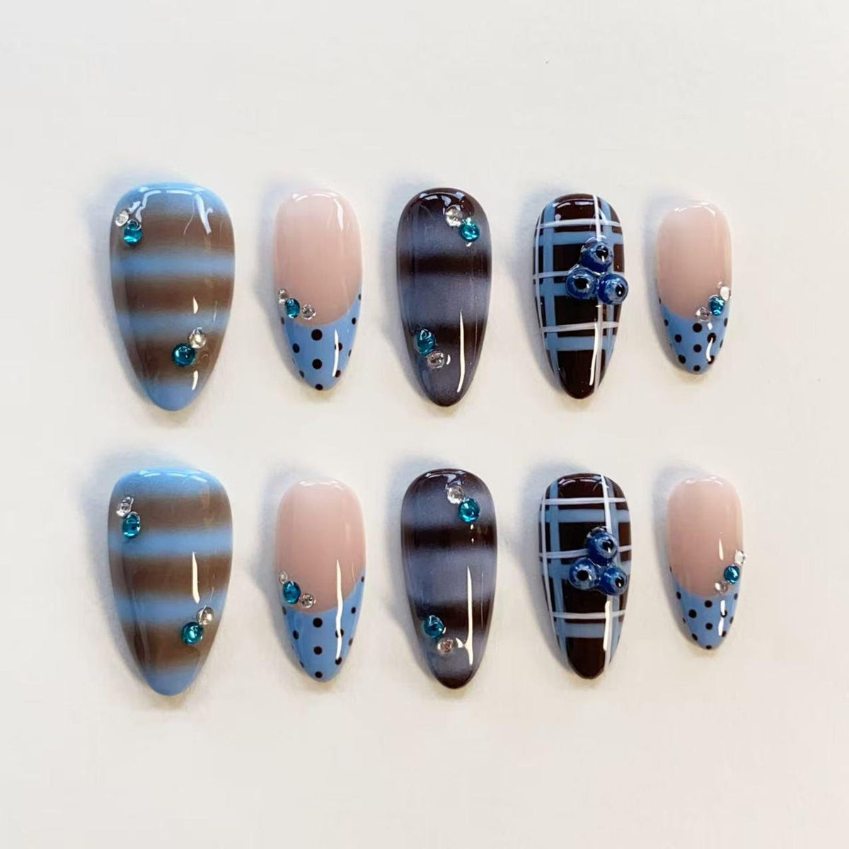 A set of ten press-on nails featuring almond-shaped tips with varied designs. Patterns include horizontal brown and blue stripes, light blue polka dots on a nude base, dark brown and blue plaid, and small blue rhinestones.