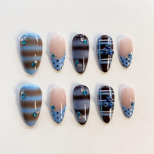 A set of ten press-on nails featuring almond-shaped tips with varied designs. Patterns include horizontal brown and blue stripes, light blue polka dots on a nude base, dark brown and blue plaid, and small blue rhinestones.