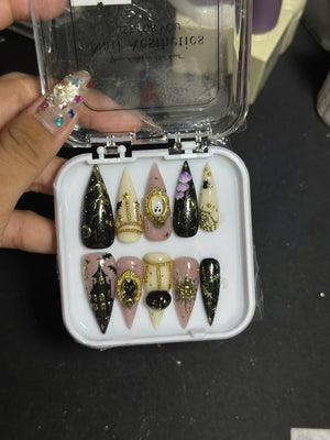 A set of ten stiletto-shaped press-on nails in a clear plastic case. The nails feature black, cream, and dusty rose base colors with intricate gold charms, metallic chains, painted haunted houses, ghosts, cats, and celestial motifs.