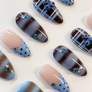 A set of almond-shaped press-on nails featuring various designs including brown and light blue plaid patterns, dark brown cat-eye effects, and light blue tips with dark brown polka dots, all accented with small teal and clear rhinestones.