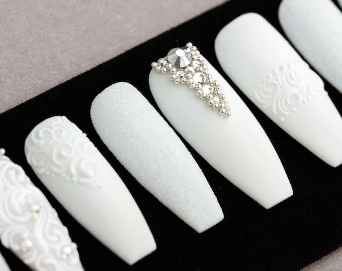 Set of ten white press-on nails with different designs. Some nails have a white glitter finish, some have a white swirl design, and some have a rhinestone design.