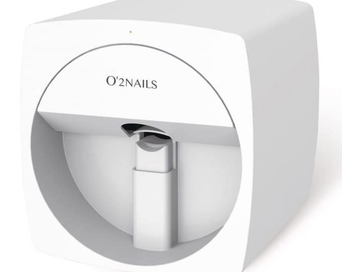 A white O'2NAILS nail art printer. The device has a square shape with rounded corners and a curved front panel. The brand name is printed on the front. The printer has an opening for nail insertion an