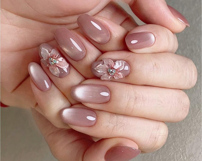 Close-up of a hand with nude-colored oval-shaped nails decorated with delicate 3D floral embellishments. The flowers are white with pink accents.