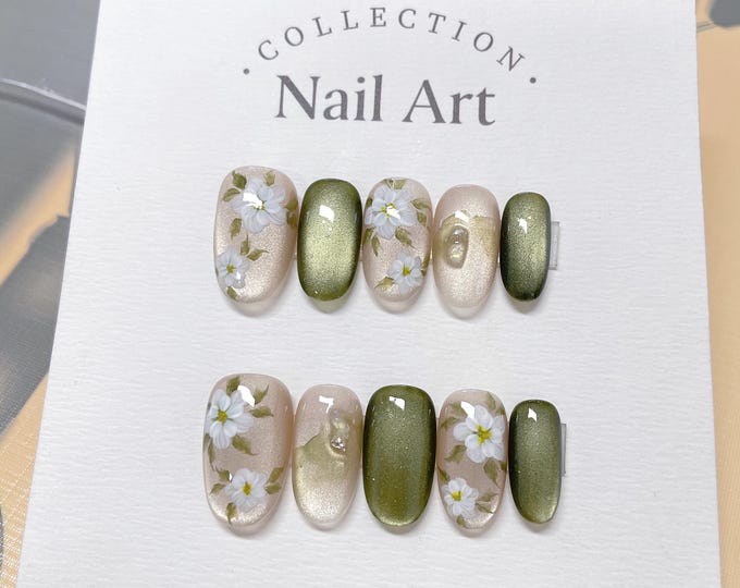 A set of six oval-shaped fake nails with a floral design. The nails are painted in shades of green and beige with white flowers and green leaves. The text "COLLECTION. Nail Art." is printed on the whi