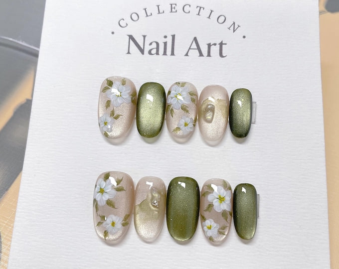 A set of six oval-shaped fake nails with a floral design. The nails are painted in shades of green and beige with white flowers and green leaves. The text "COLLECTION. Nail Art." is printed on the whi