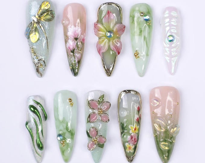 Ten long, almond-shaped artificial nails with various floral and nature-inspired designs. The designs include a dragonfly, flowers, leaves, and swirls. Some nails have rhinestones and gold accents.